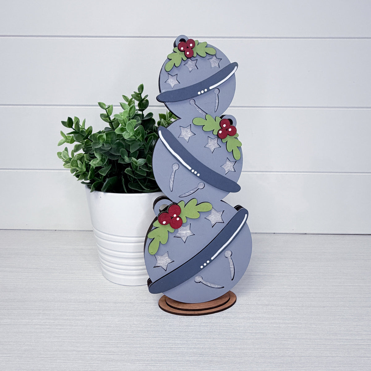 A decorative stacked ornament made from gray wood, decorated with green pine branches, red berries, and white stars, displayed on a wooden stand next to a potted green plant against a white paneled background.