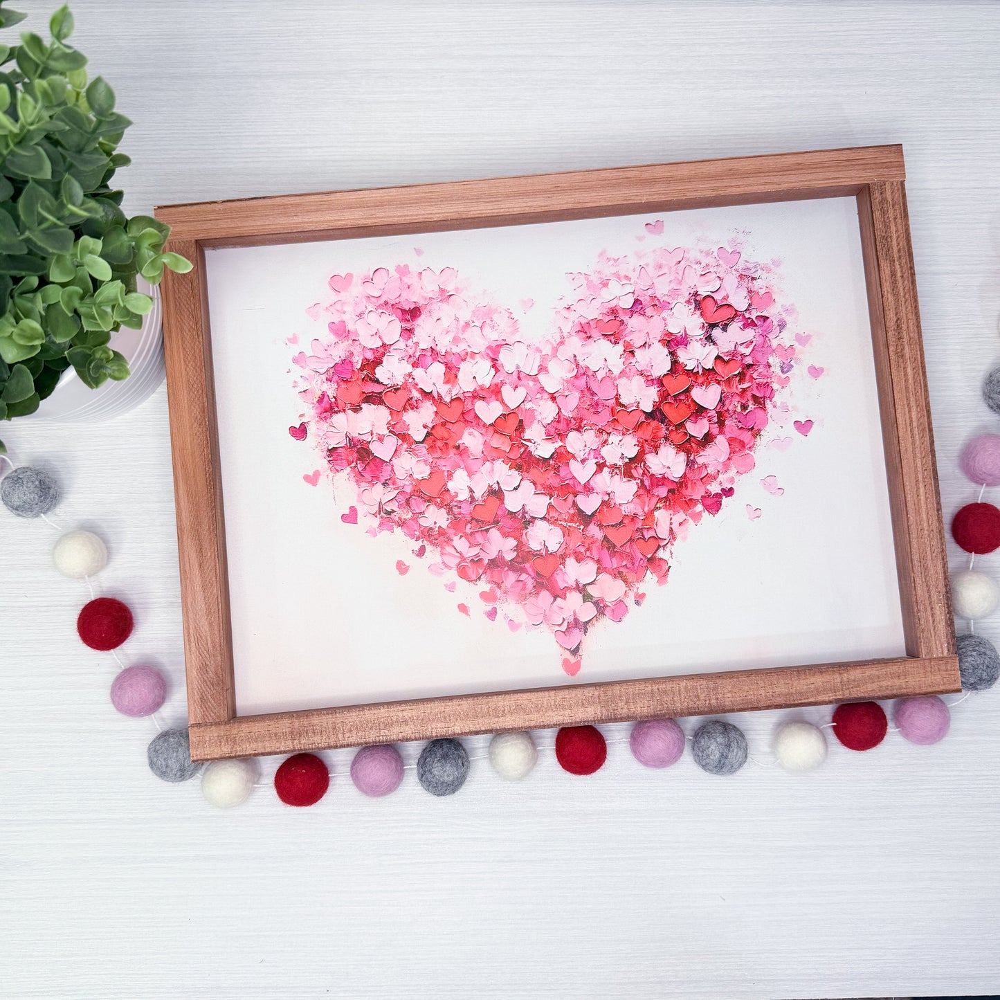 Heart-shaped arrangement of pink and red hearts in a wooden frame on a white surface with decorative pom-poms and a plant.
