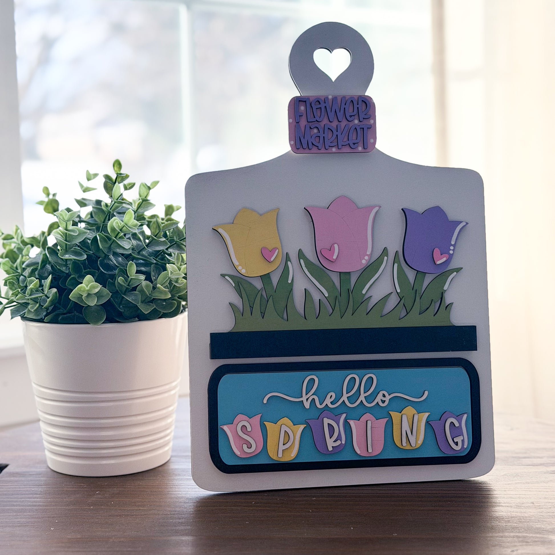 Decorative sign with tulips and 'hello spring' text on a wooden surface with a plant in the background.