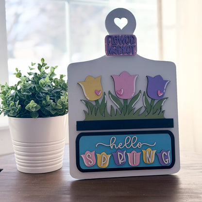 Decorative sign with tulips and 'hello spring' text on a wooden surface with a plant in the background.