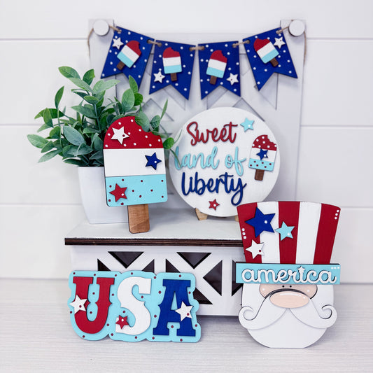 Decorative items with patriotic theme including a gnome, 'USA' sign, and 'Sweet Land of Liberty' plate on a white background.