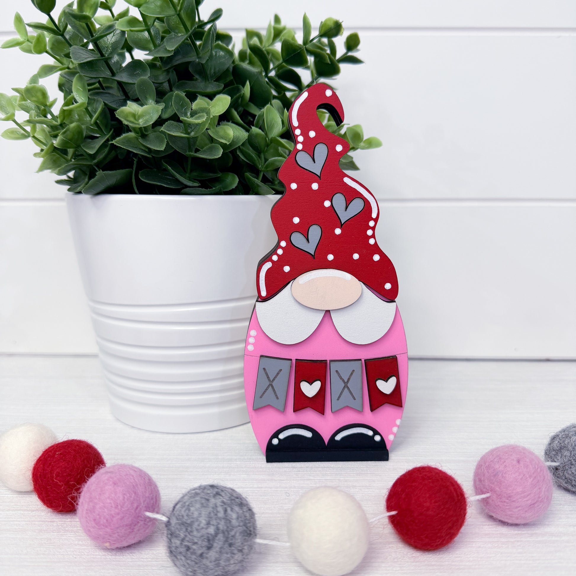 Decorative gnome with red hat and pink body next to a potted plant and colorful pom-pom garland.