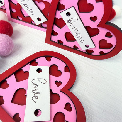 Heart-shaped pink and red cutout decorations with 'Love' and 'Be mine' tags on a white background.