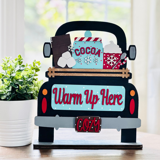 A decorative wood truck with interchangeable inserts that reads 'Warm Up Here' with a cocoa mug and snowflake design, placed on a table.