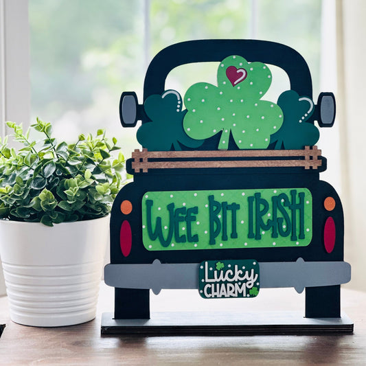 Decorative sign of a truck with shamrocks and text on a wooden surface with a plant in the background.