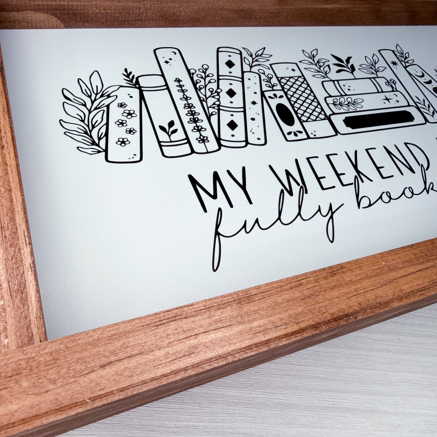 Wooden frame with a white background featuring black line art of books and text 'My weekend fully book'.