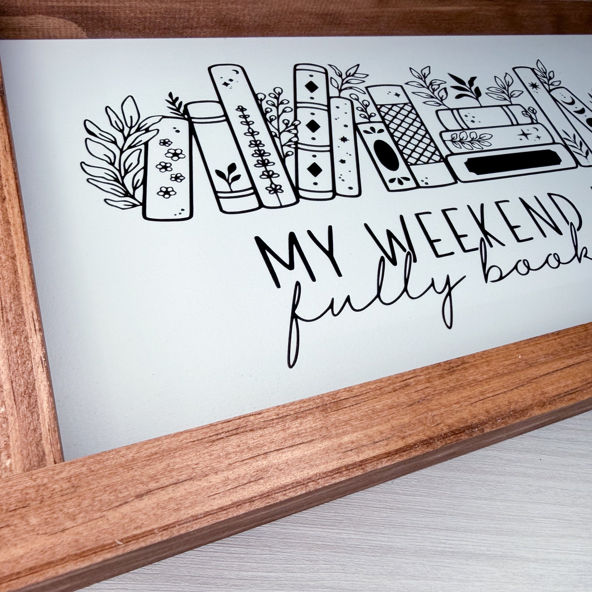 Wooden frame with a white background featuring black line art of books and text 'My weekend fully book'.