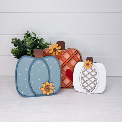 Three decorative layered wooden pumpkins arranged on a light wood surface against a white shiplap wall. The left pumpkin is dusty blue with white dots and a yellow flower; the center pumpkin is orange-brown with a lattice pattern and a red heart; the right pumpkin is white with a lattice pattern and a yellow flower.