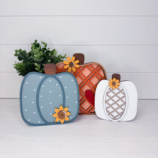Three decorative layered wooden pumpkins arranged on a light wood surface against a white shiplap wall. The left pumpkin is dusty blue with white dots and a yellow flower; the center pumpkin is orange-brown with a lattice pattern and a red heart; the right pumpkin is white with a lattice pattern and a yellow flower.
