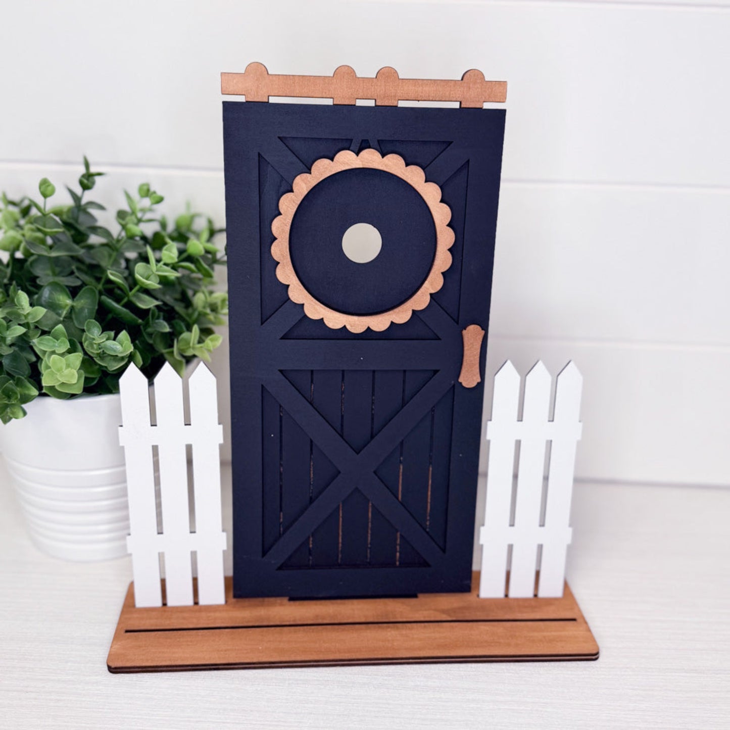 Decorative barn door with floral design on a stand, surrounded by plants and white decor.