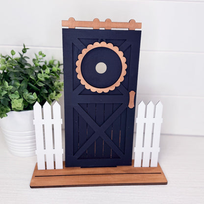 Decorative barn door with floral design on a stand, surrounded by plants and white decor.