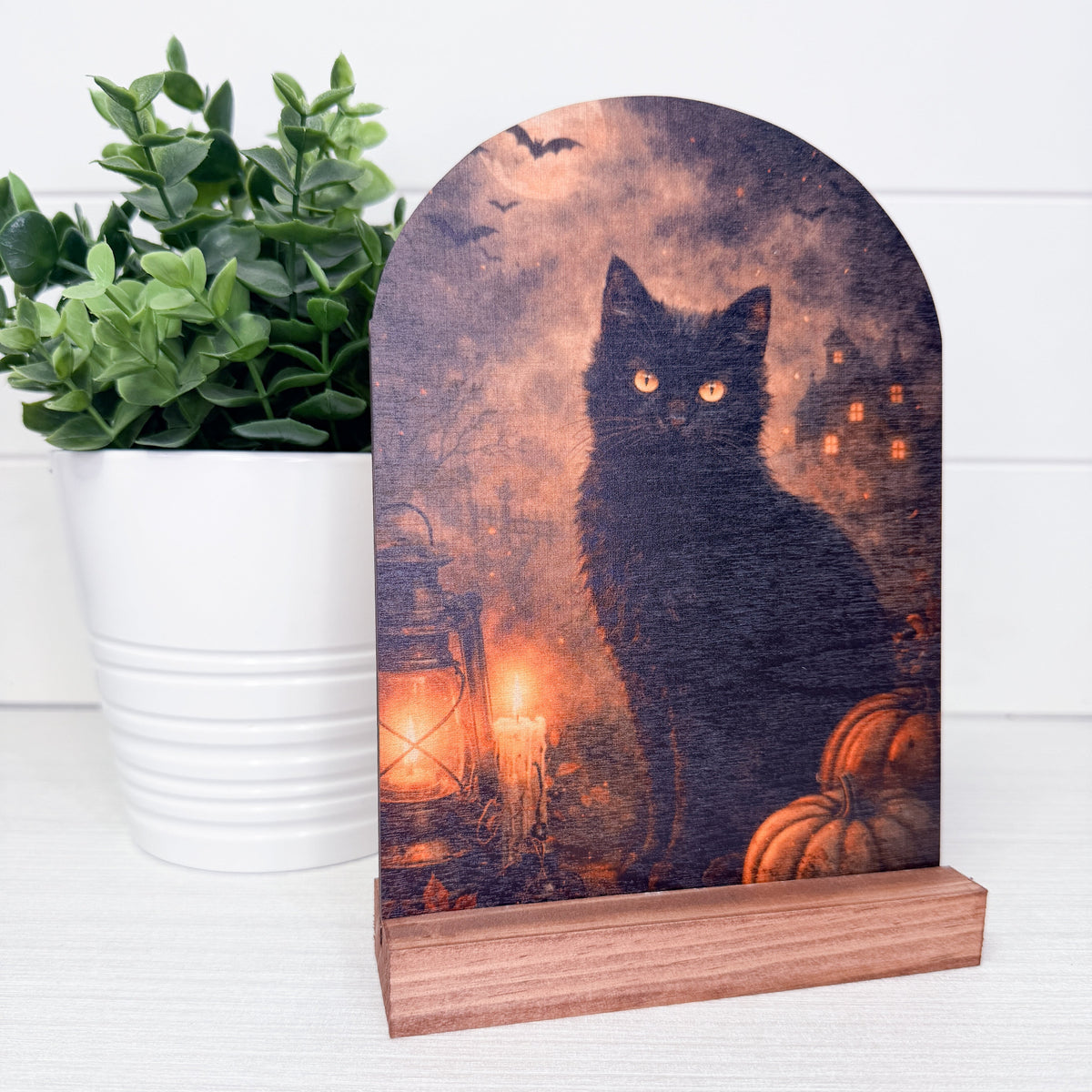 A Halloween-themed wooden plaque featuring a black cat silhouette against a spooky haunted-house night scene with orange lanterns and pumpkins, displayed on a wooden base next to a potted green plant.