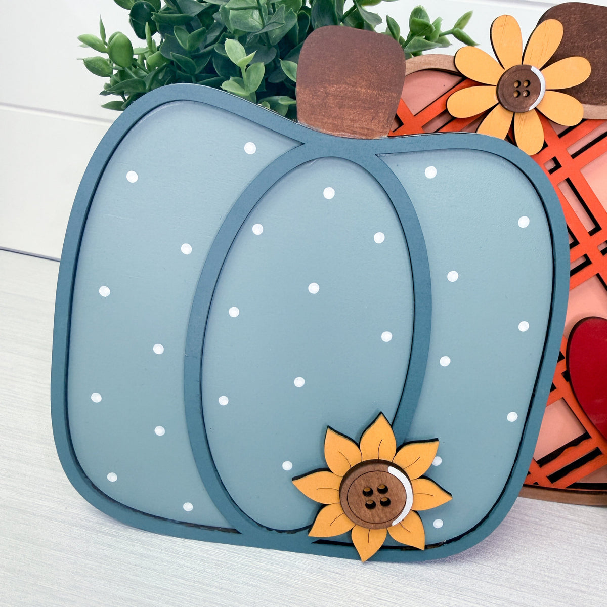 A decorative autumn pumpkin craft piece in dusty blue with white polka dots, a brown wooden stem, and a yellow sunflower embellishment, displayed on a white surface with other fall-themed decor in the background.