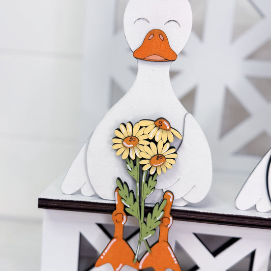 A decorative wooden duck figurine holding yellow daisies with green stems, shown close up against a white background.