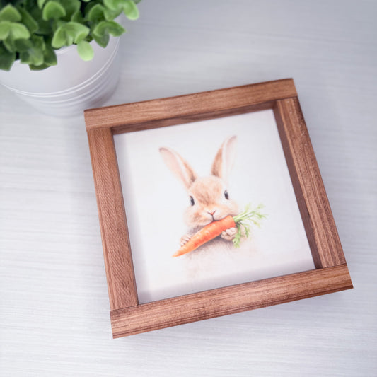 Framed artwork of a rabbit holding a carrot on a white surface with a plant in the background.
