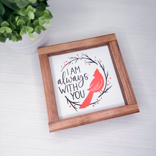 Framed artwork with a red cardinal and text on a white background, placed on a light surface with a plant in the corner.