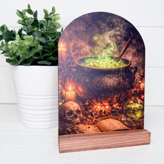 A decorative tabletop sign shaped like a tombstone, printed with a spooky cauldron scene and mounted on a wooden base, styled on a white surface next to a potted green plant.
