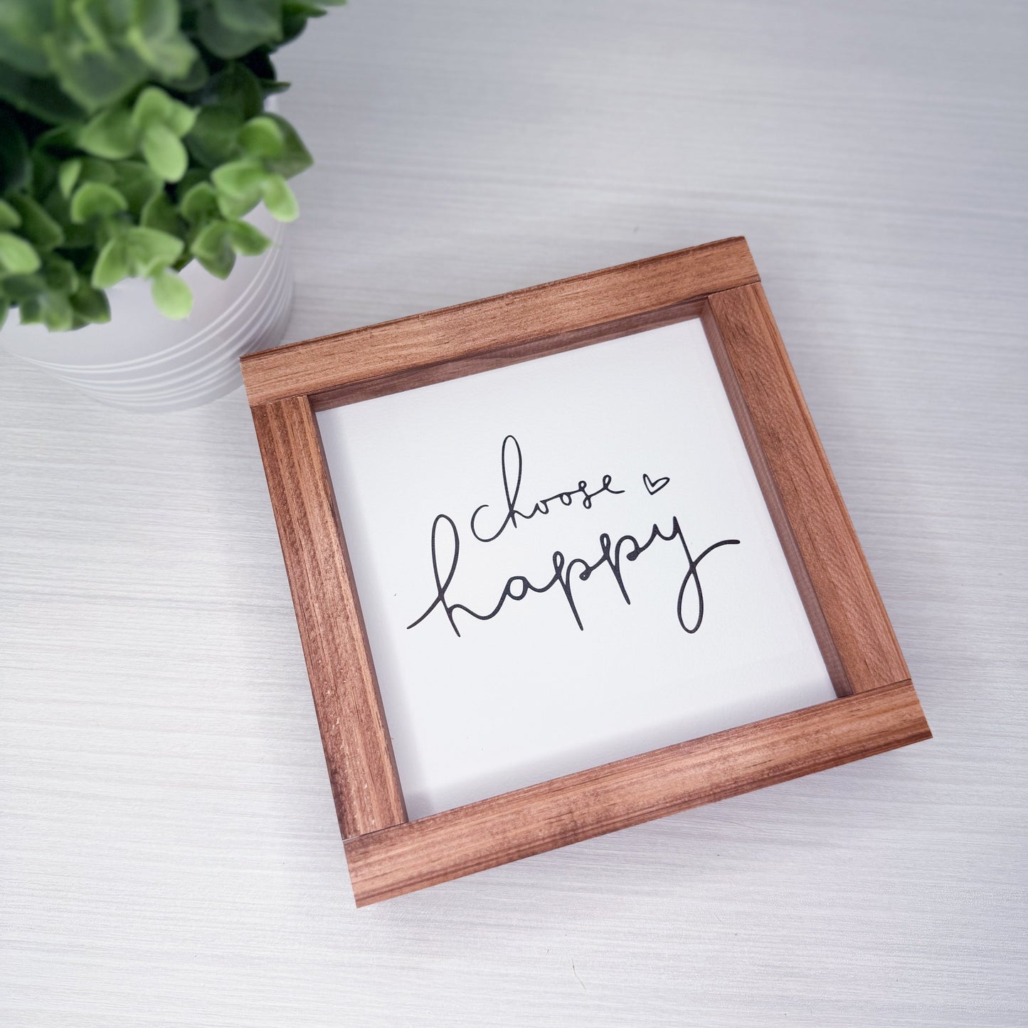 Wooden-framed sign with 'Choose Happy' text on a white surface with a plant in the corner.