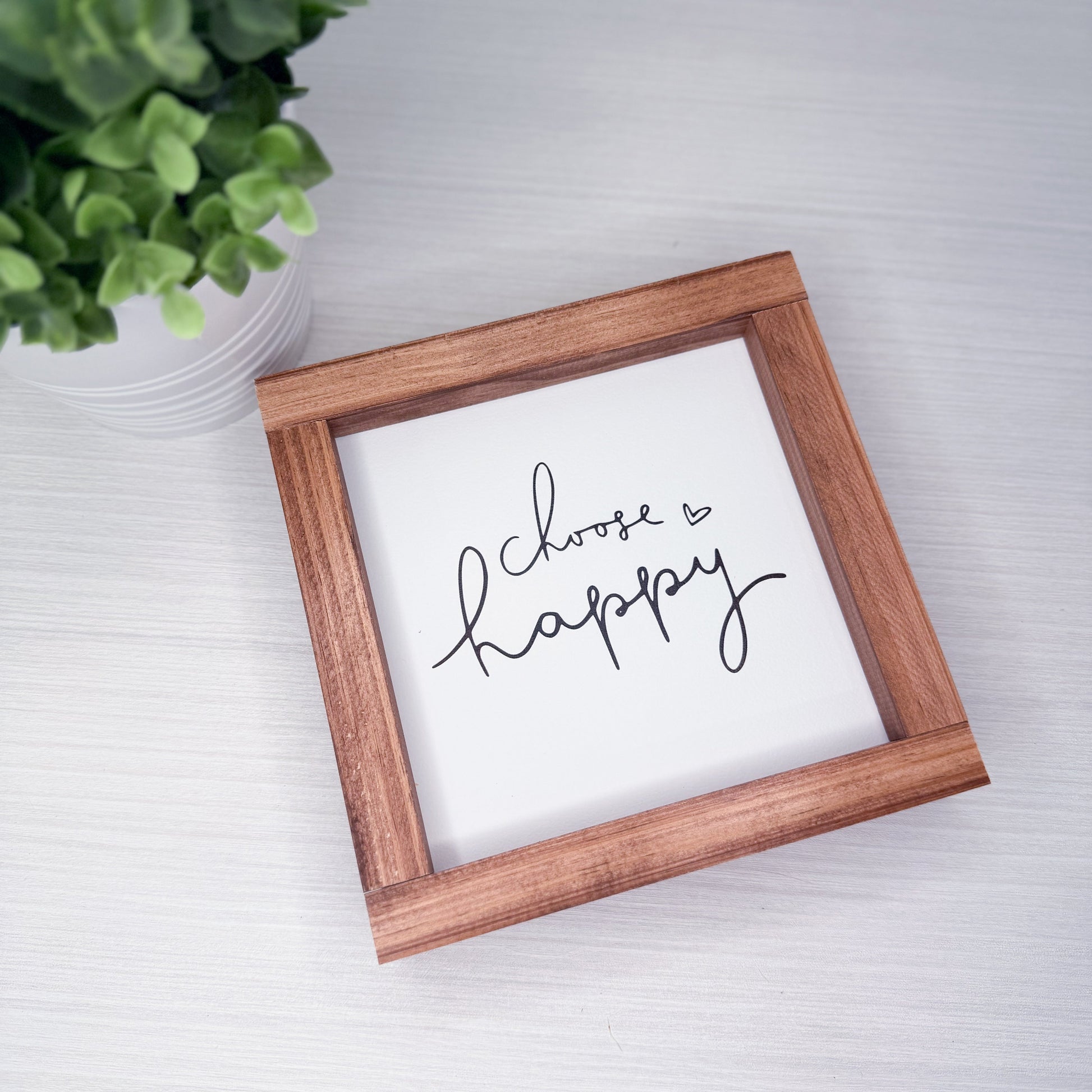 Wooden-framed sign with 'Choose Happy' text on a white surface with a plant in the corner.