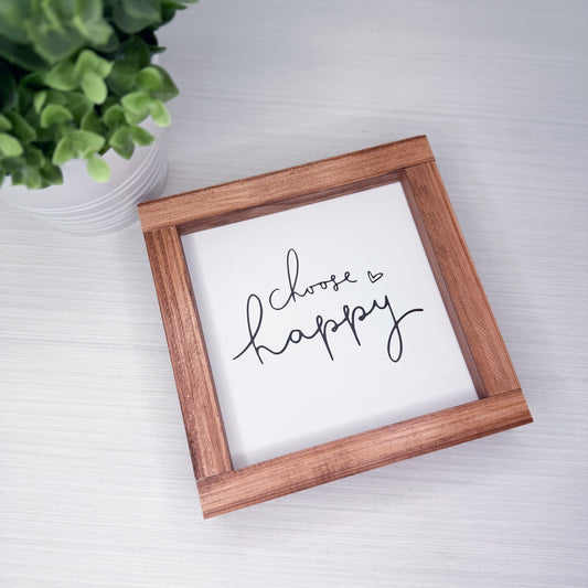 Wooden-framed sign with 'Choose Happy' text on a white surface with a plant in the corner.