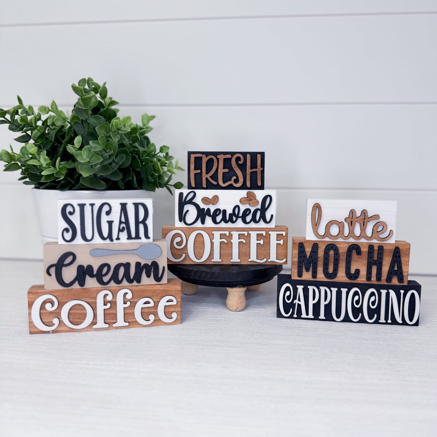 Set of wooden signs with coffee-themed text on a white surface with a plant in the background.
