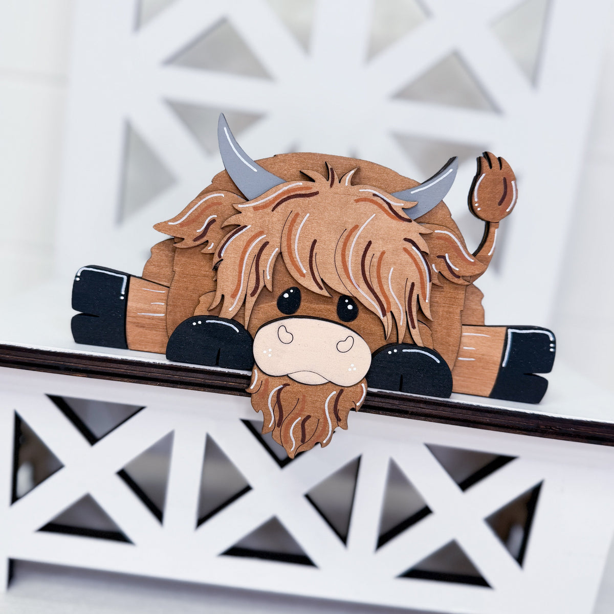 A wooden Highland cow wall decor piece with a shaggy brown faux-fur design, gray horns, and black hooves, shown leaning on a white decorative lattice shelf.