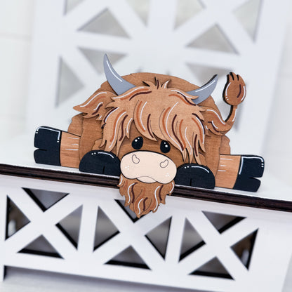 A wooden Highland cow wall decor piece with a shaggy brown faux-fur design, gray horns, and black hooves, shown leaning on a white decorative lattice shelf.