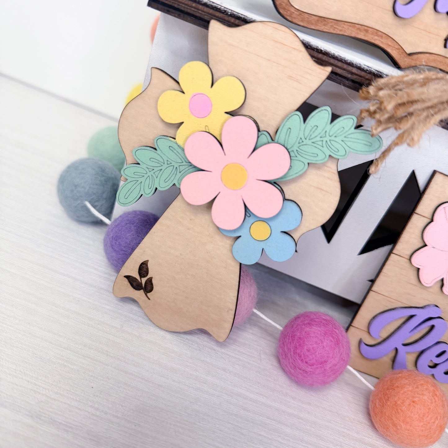 Decorative wooden letters with floral designs and colorful pom-poms on a textured surface.