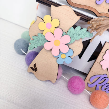 Decorative wooden letters with floral designs and colorful pom-poms on a textured surface.