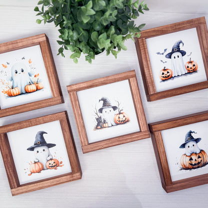 Five small framed Halloween art prints arranged on a light wood surface with a green plant above. The prints show cute ghost and pumpkin illustrations in different poses, each in a brown wooden frame.