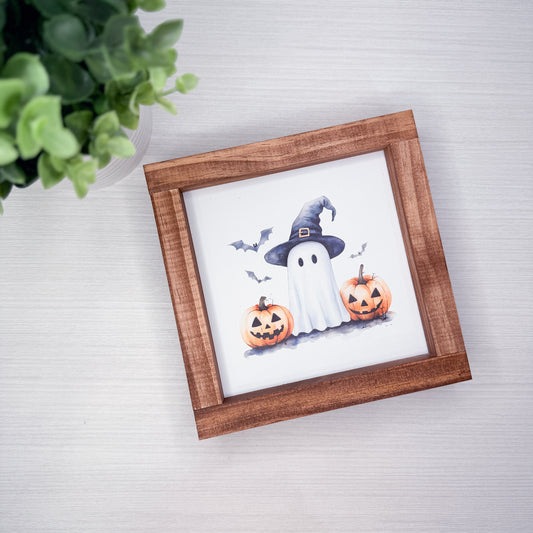 A square framed Halloween art print featuring a white ghost in a black witch hat with two orange jack-o'-lantern pumpkins and small bats, displayed on a light gray wood-textured surface next to a green plant.