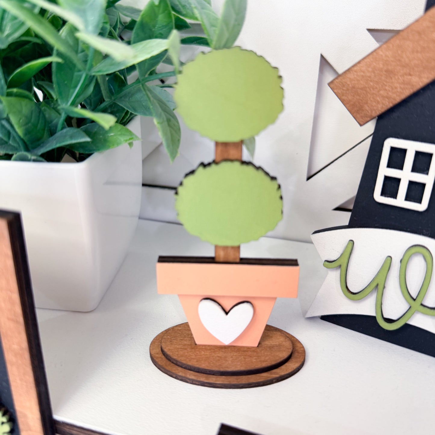 Decorative topiary with green balls on a wooden stand, placed on a surface with a house-shaped object and plant in the background.