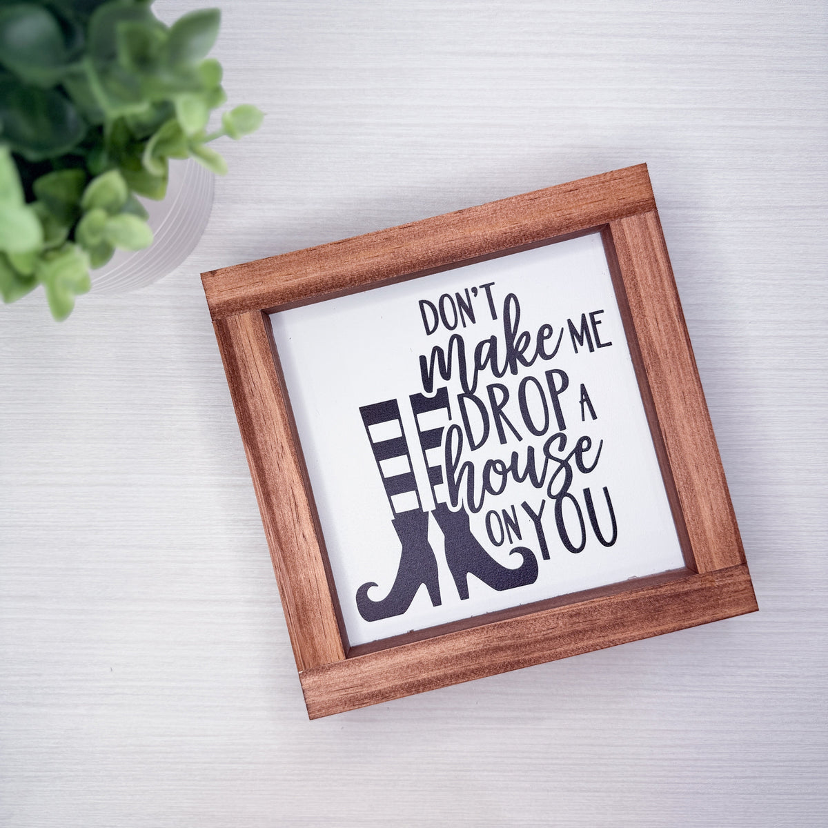A square framed decorative sign with the phrase “DON’T make me DROP a house ON YOU” in black script and block lettering over a white background, with a black-and-white striped boot graphic. The sign is shown in a medium brown wooden frame on a light gray surface, with a small green plant partially visible in the top-left corner.