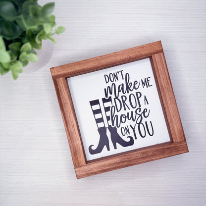 A square framed decorative sign with the phrase “DON’T make me DROP a house ON YOU” in black script and block lettering over a white background, with a black-and-white striped boot graphic. The sign is shown in a medium brown wooden frame on a light gray surface, with a small green plant partially visible in the top-left corner.