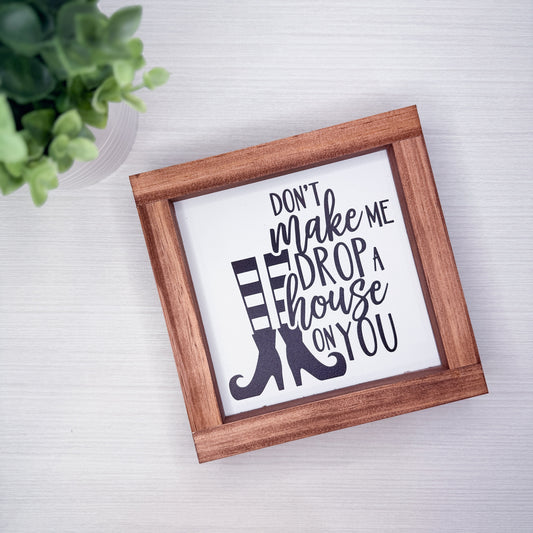A square framed decorative sign with the phrase “DON’T make me DROP a house ON YOU” in black script and block lettering over a white background, with a black-and-white striped boot graphic. The sign is shown in a medium brown wooden frame on a light gray surface, with a small green plant partially visible in the top-left corner.