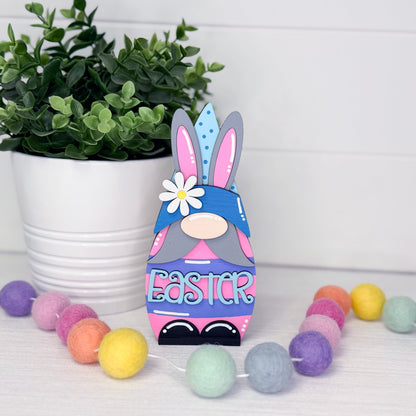 Colorful Easter gnome with bunny ears next to a potted plant and decorative eggs on a white surface.