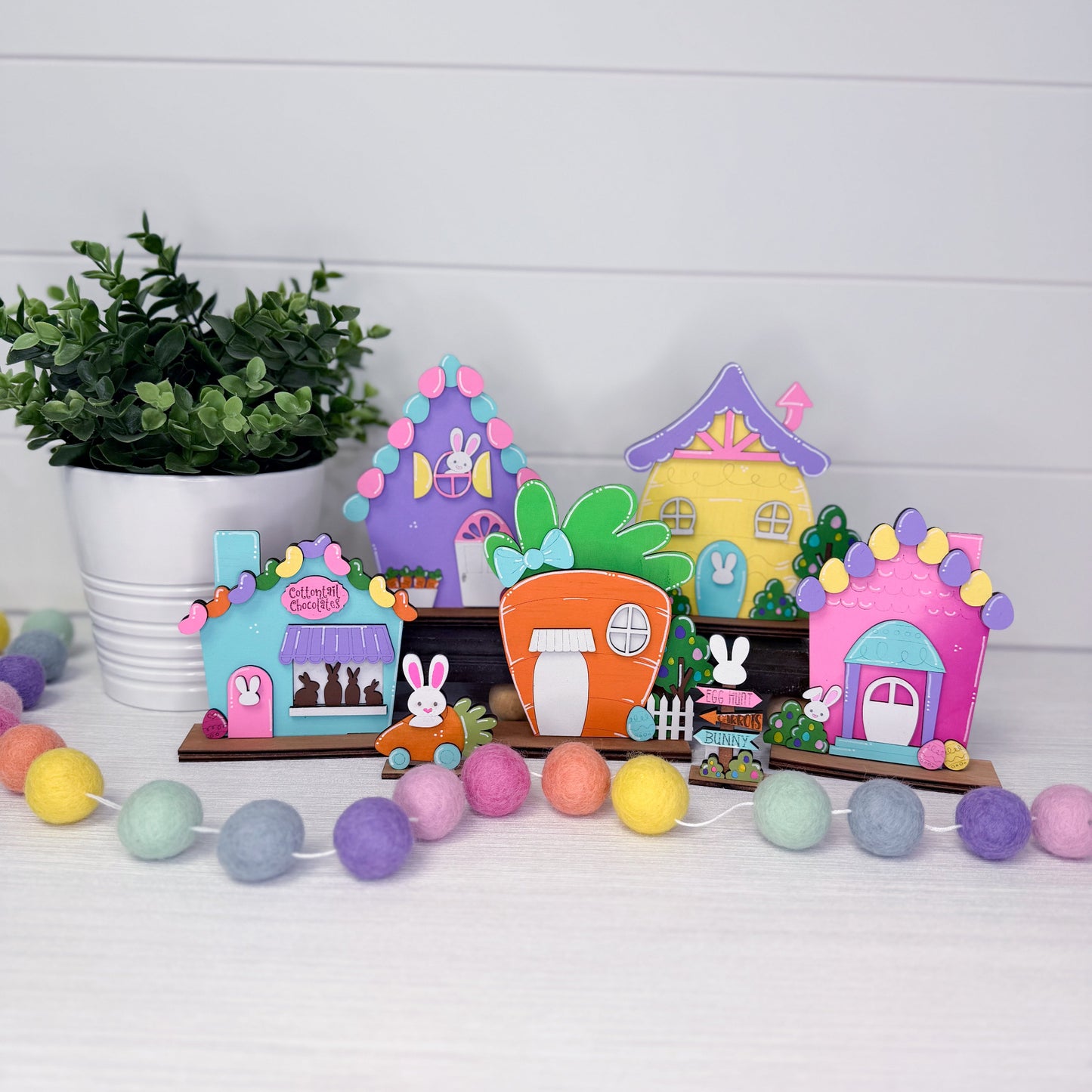 Colorful miniature houses with decorative elements on a white surface with a plant and string lights in the background.