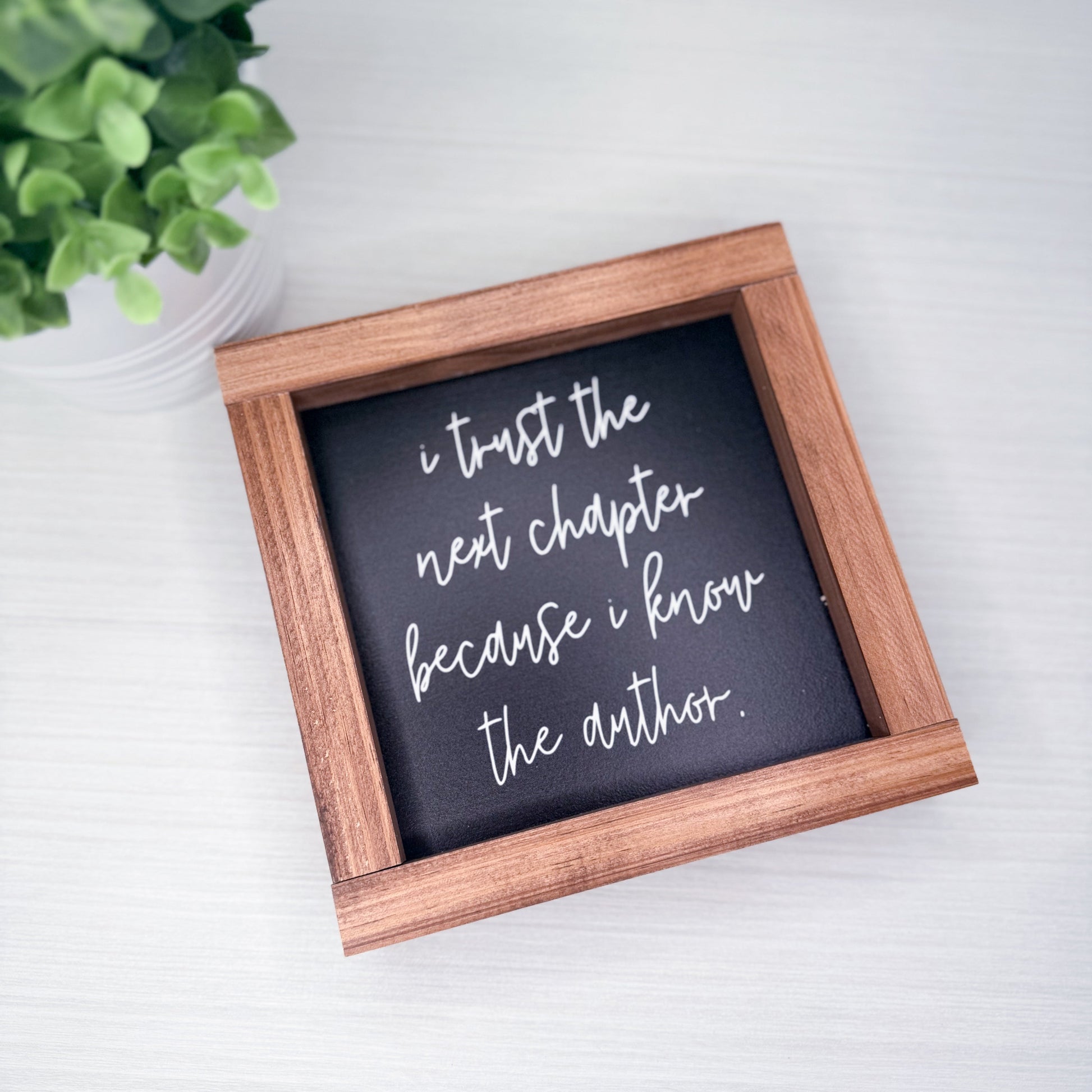 Wooden-framed sign with motivational quote on a white surface with a plant in the background