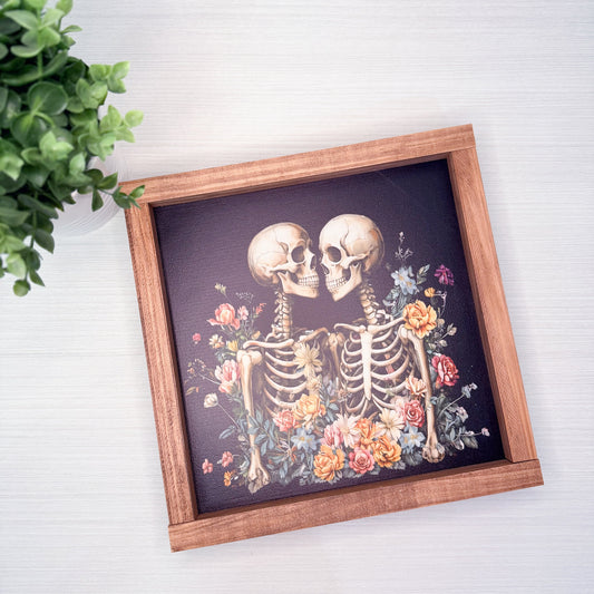 Framed artwork of two kissing skeletons surrounded by flowers on a white surface with a plant in the corner.