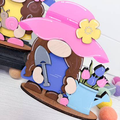 Colorful wooden gnome figurine with a pink hat and garden tools on a white surface.