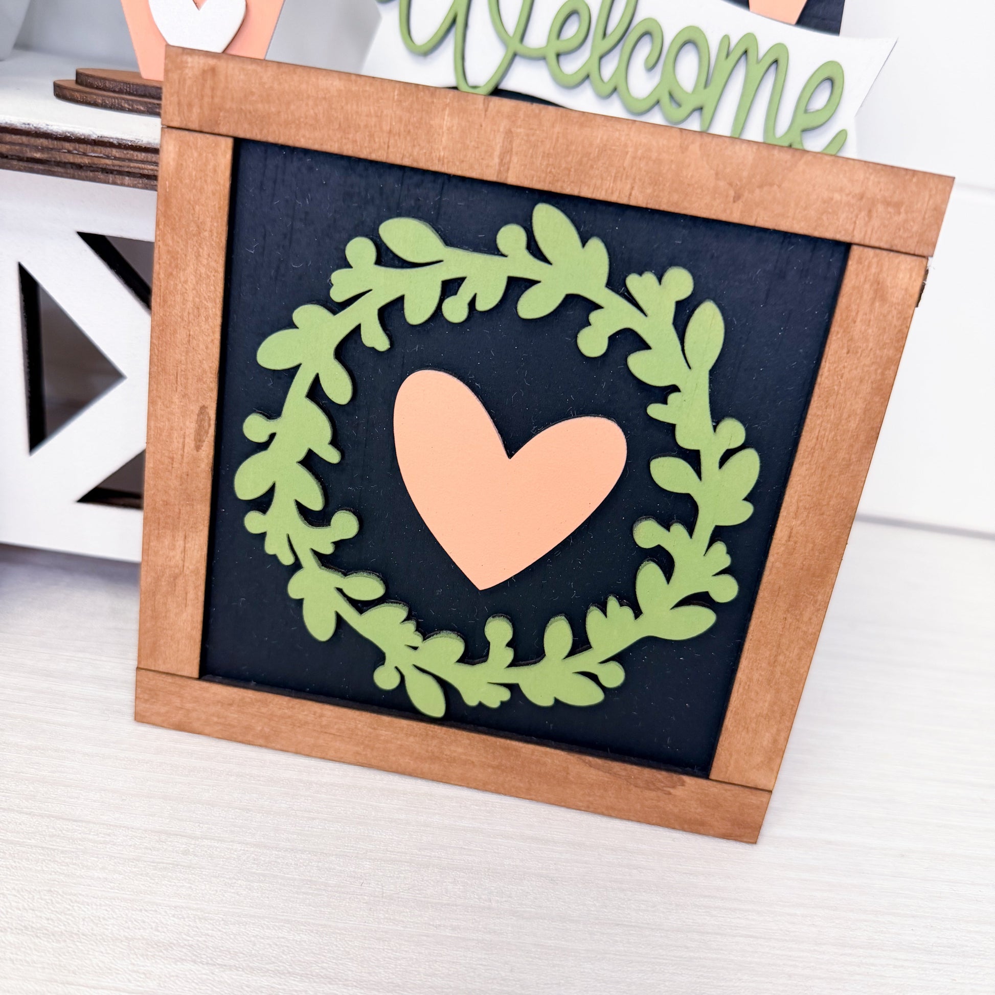 Decorative sign with a heart and leaf wreath design in a wooden frame.