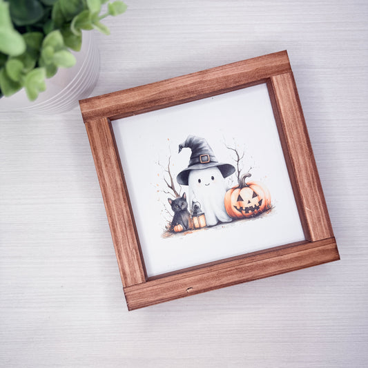 A square wooden picture frame with a white mat, featuring a cute Halloween illustration of a ghost, a black cat, and a jack-o'-lantern pumpkin, displayed on a light gray wood-textured surface next to a green plant.