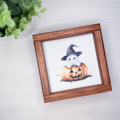 A square wooden picture frame with a watercolor illustration of a cute white ghost wearing a witch hat sitting on a carved orange pumpkin, displayed on a light gray wood-textured surface next to a green plant.