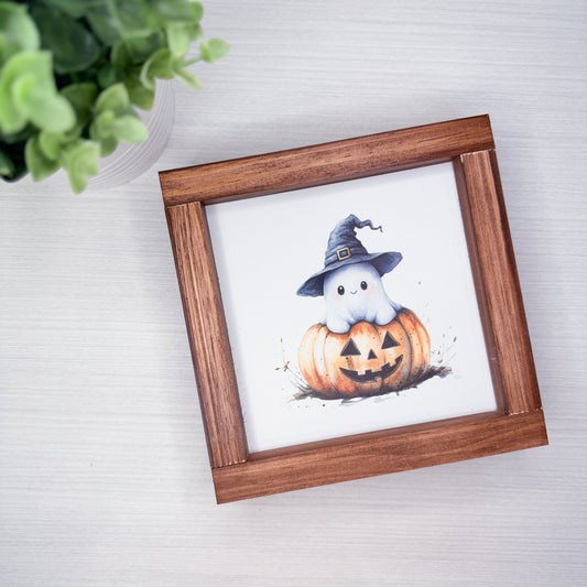 A square wooden picture frame with a watercolor illustration of a cute white ghost wearing a witch hat sitting on a carved orange pumpkin, displayed on a light gray wood-textured surface next to a green plant.