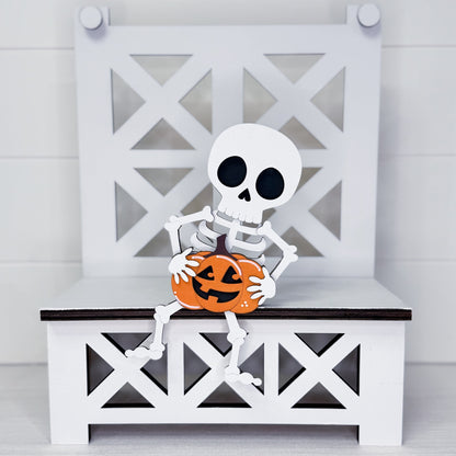 A decorative white skeleton figurine sitting on a white bench with a carved orange pumpkin, set against a white paneled background.