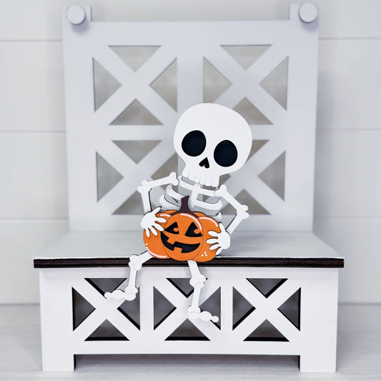 A decorative white skeleton figurine sitting on a white bench with a carved orange pumpkin, set against a white paneled background.