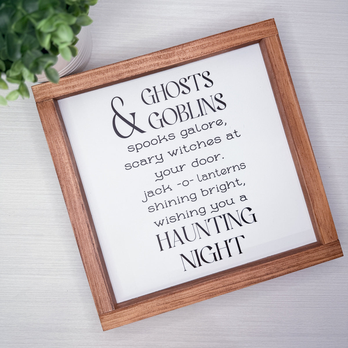 A framed typographic wall sign with a wooden brown frame and white background, printed with the phrase “Ghosts & Goblins / spooks galore, scary witches at your door / jack -o- lanterns shining bright, wishing you a HAUNTING NIGHT.” The sign is photographed on a light wood surface with a small green plant partially visible in the top left.
