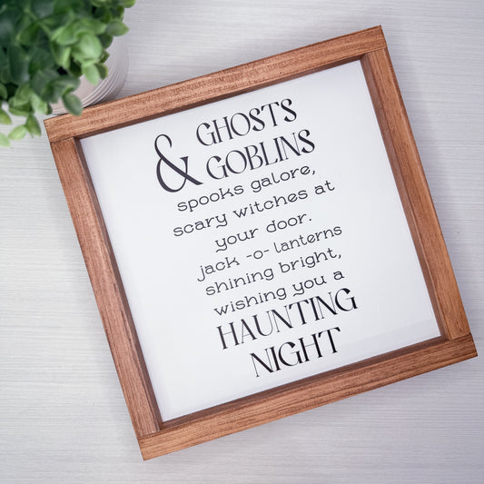 A framed typographic wall sign with a wooden brown frame and white background, printed with the phrase “Ghosts & Goblins / spooks galore, scary witches at your door / jack -o- lanterns shining bright, wishing you a HAUNTING NIGHT.” The sign is photographed on a light wood surface with a small green plant partially visible in the top left.