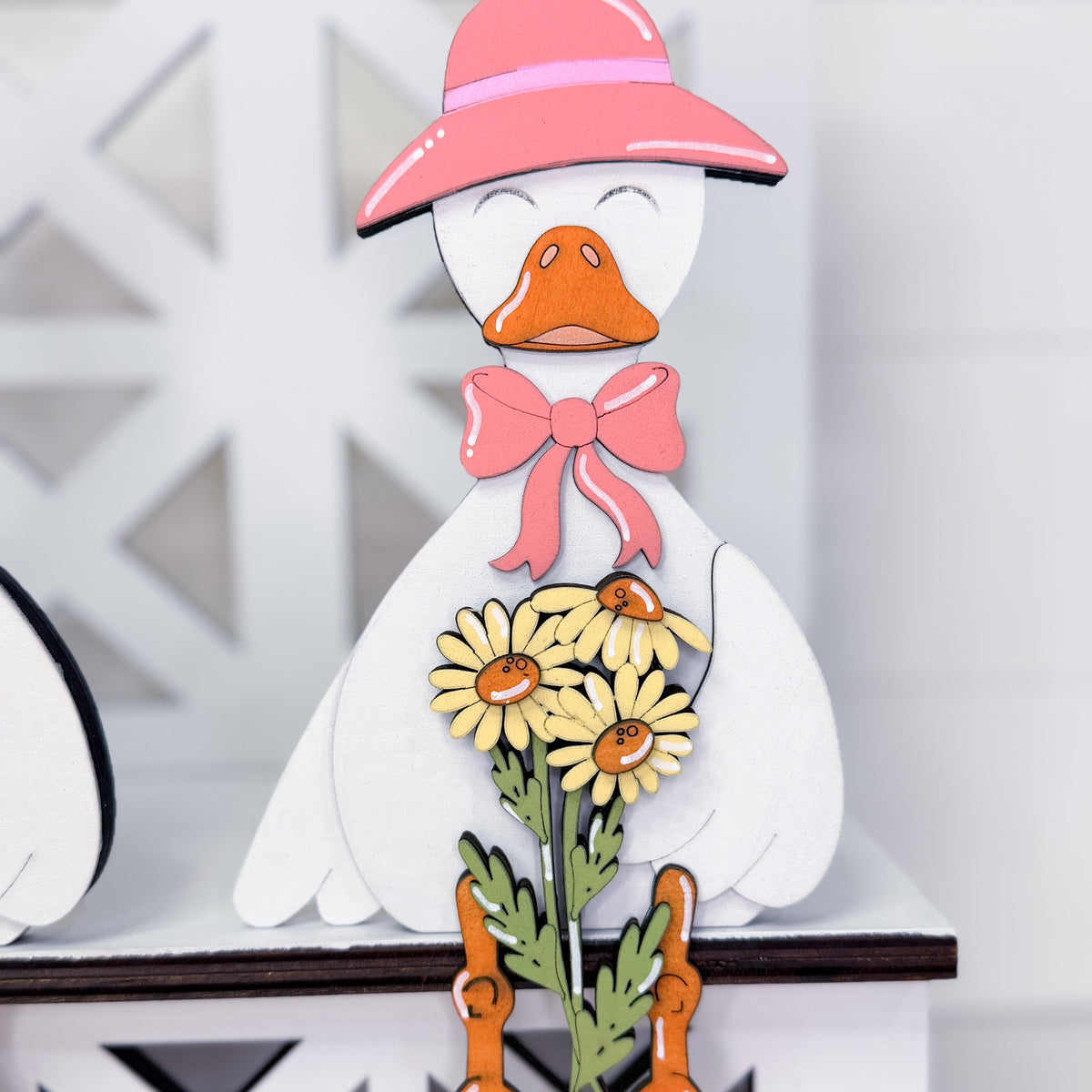 A decorative wooden duck figurine with a pink hat, pink bow, yellow daisies, and green stems, displayed on a white shelf.
