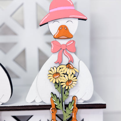 A decorative wooden duck figurine with a pink hat, pink bow, yellow daisies, and green stems, displayed on a white shelf.